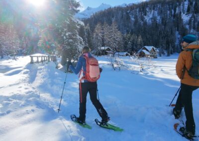Two hikers snowshoeing through a snowy alpine valley with mountain cabins and forests in Val di Pejo, Trentino