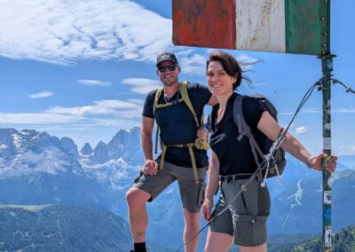 Two hikers standing beside an Italian flag on a mountain summit in the Dolomites